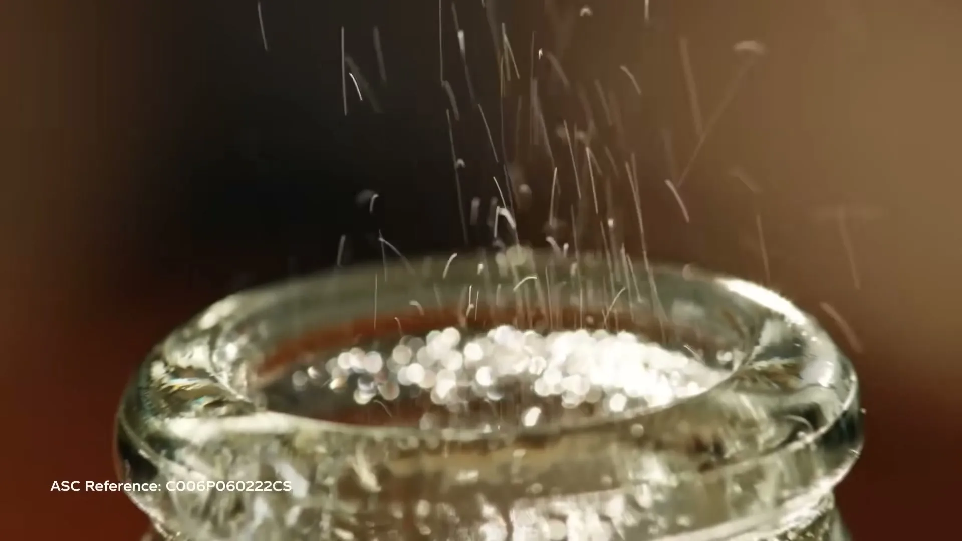 Tiny white particles fall into a clear glass bottle, creating a fizzy effect, in a close-up shot.