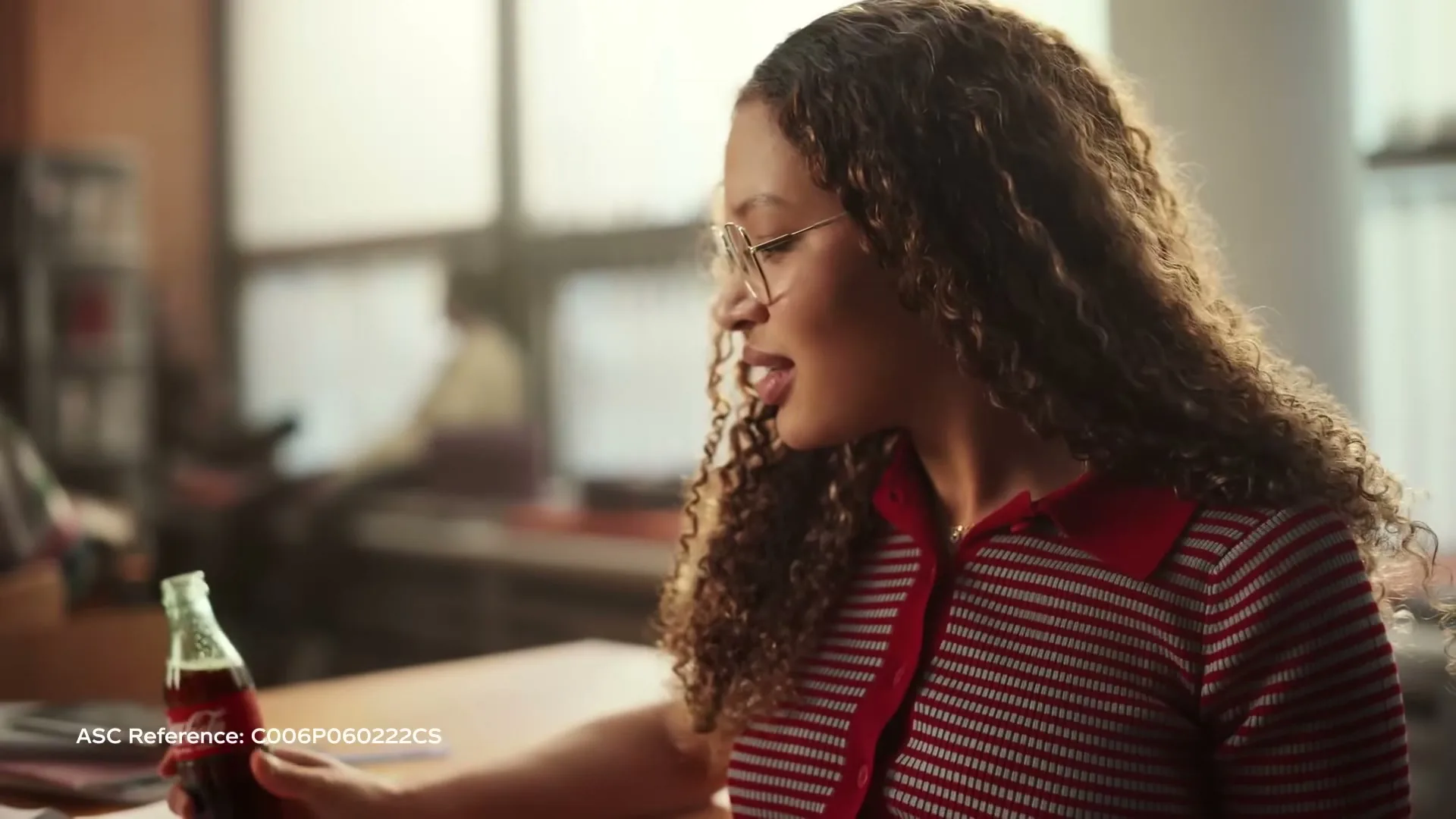 A young woman with curly hair and glasses looks down at a Coca-Cola bottle in her hand, in an office setting.