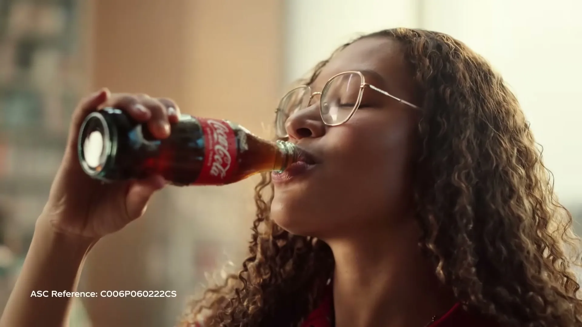 A young woman with curly hair and glasses drinks from a glass bottle of Coca-Cola, with a soft, warm light illuminating her face.