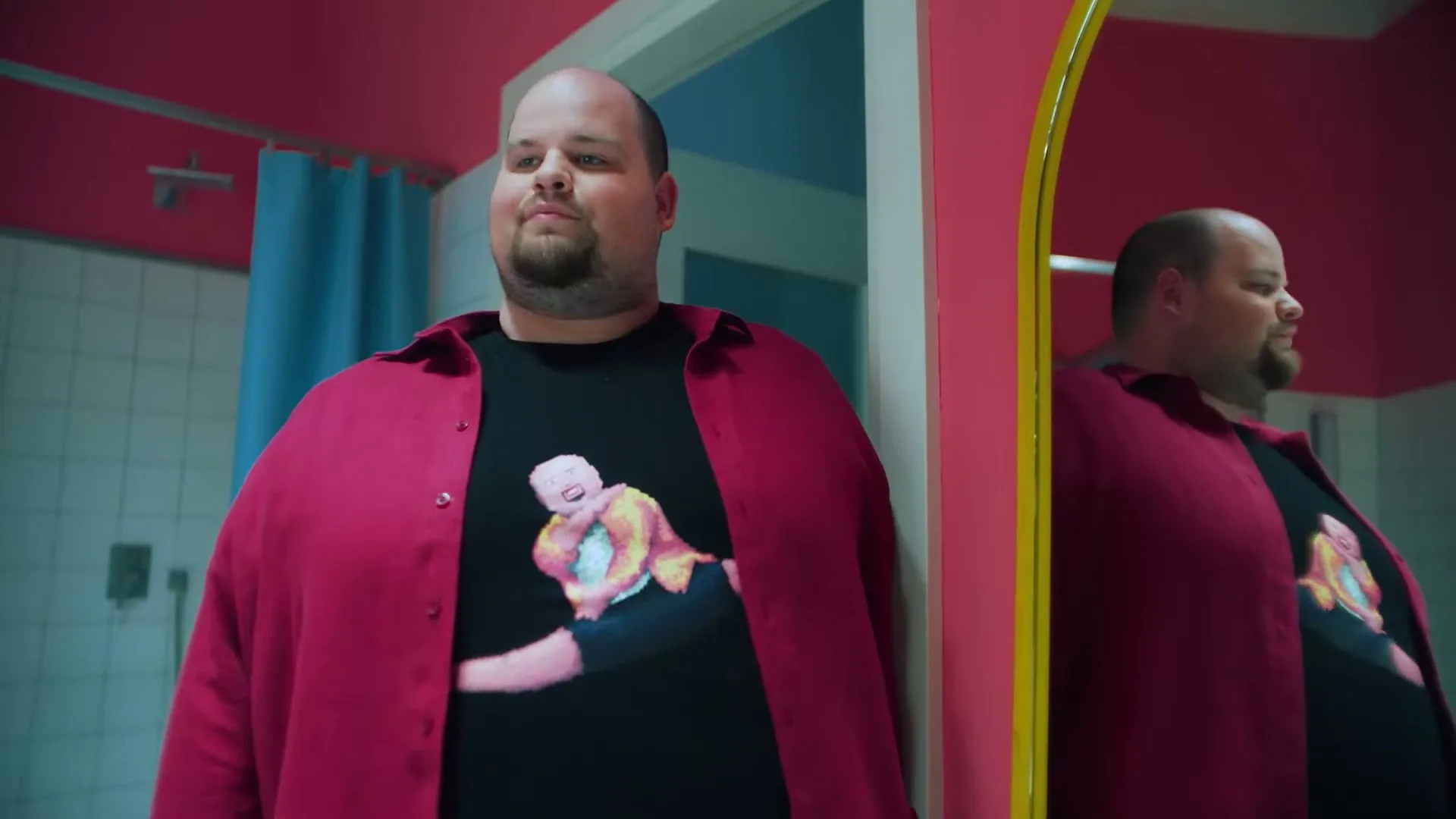 A man with a beard and a pink shirt over a black t-shirt with a pixelated dancer design looks at his reflection in a bathroom mirror.