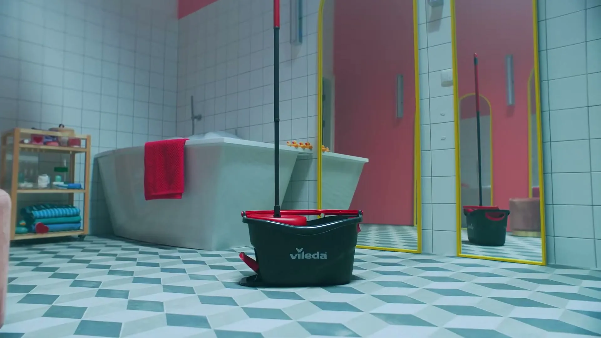 A Vileda mop and bucket stand on a tiled floor in a pink and white bathroom with a bathtub and mirrors.