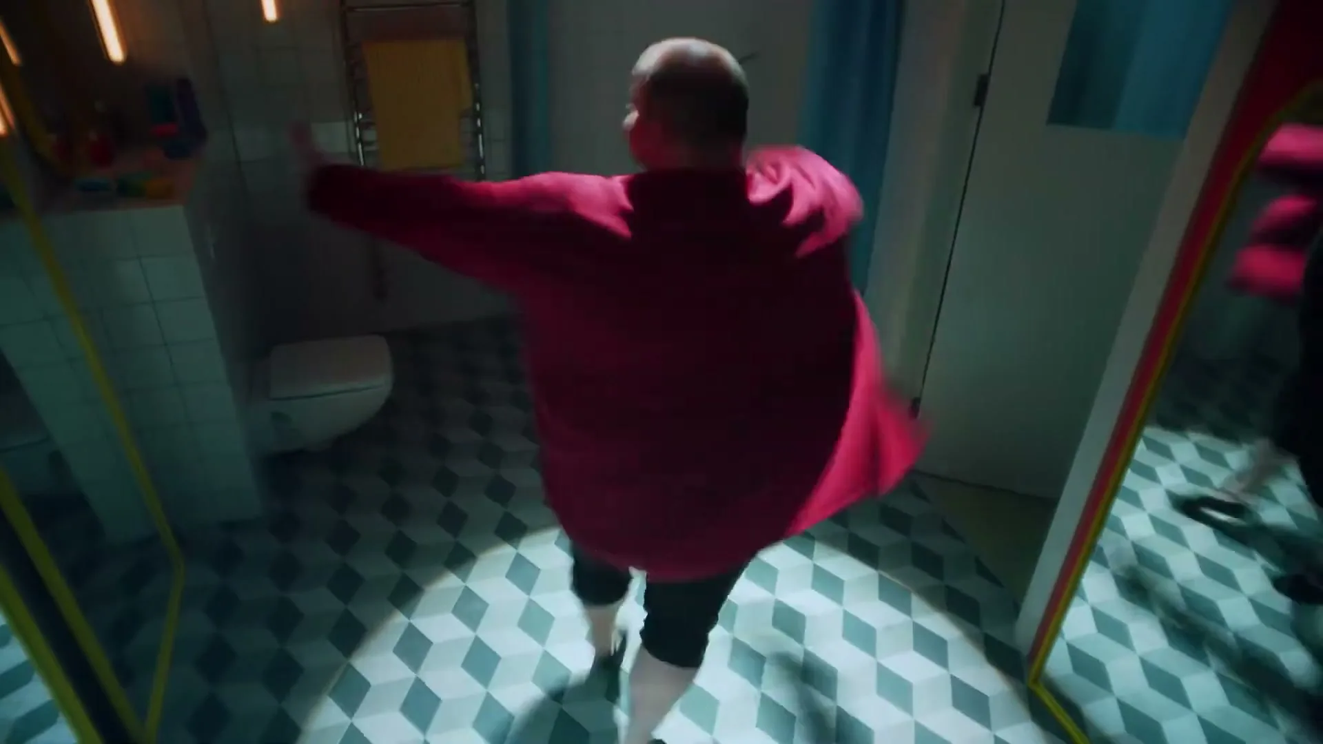 A bald man in a red shirt and black pants dances energetically in a bathroom with geometric floor tiles, his reflection visible in a mirror.