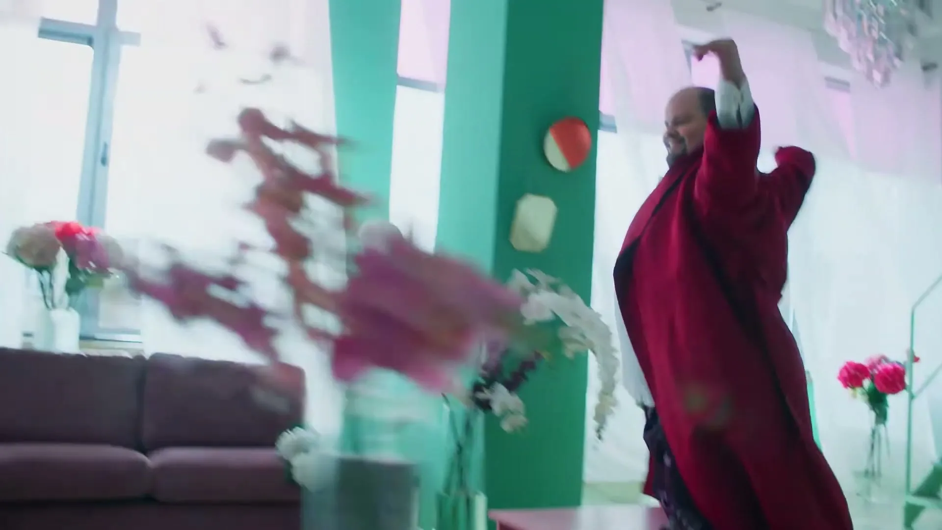 A man in a red coat and white shirt dances joyfully on a pink wheeled table in a brightly lit, colorful living room.
