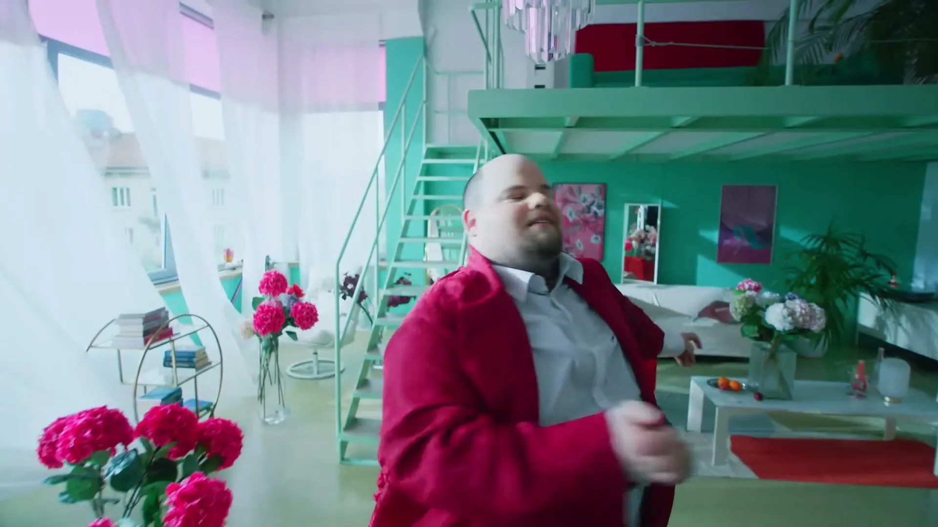 A man in a red jacket and white shirt dances energetically in a brightly lit, colorful apartment with a loft bed and sheer curtains.