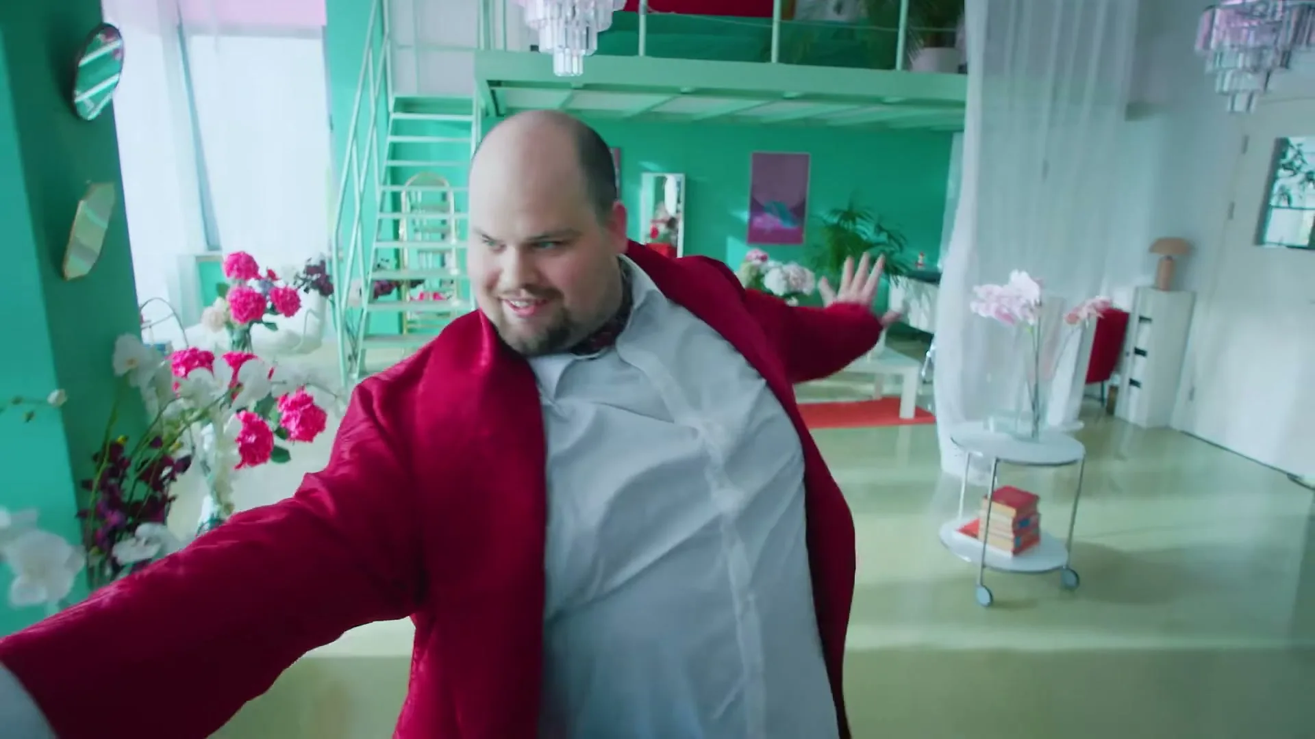 A bald man in a red robe and white shirt dances energetically in a brightly lit, colorful apartment with a mezzanine level.
