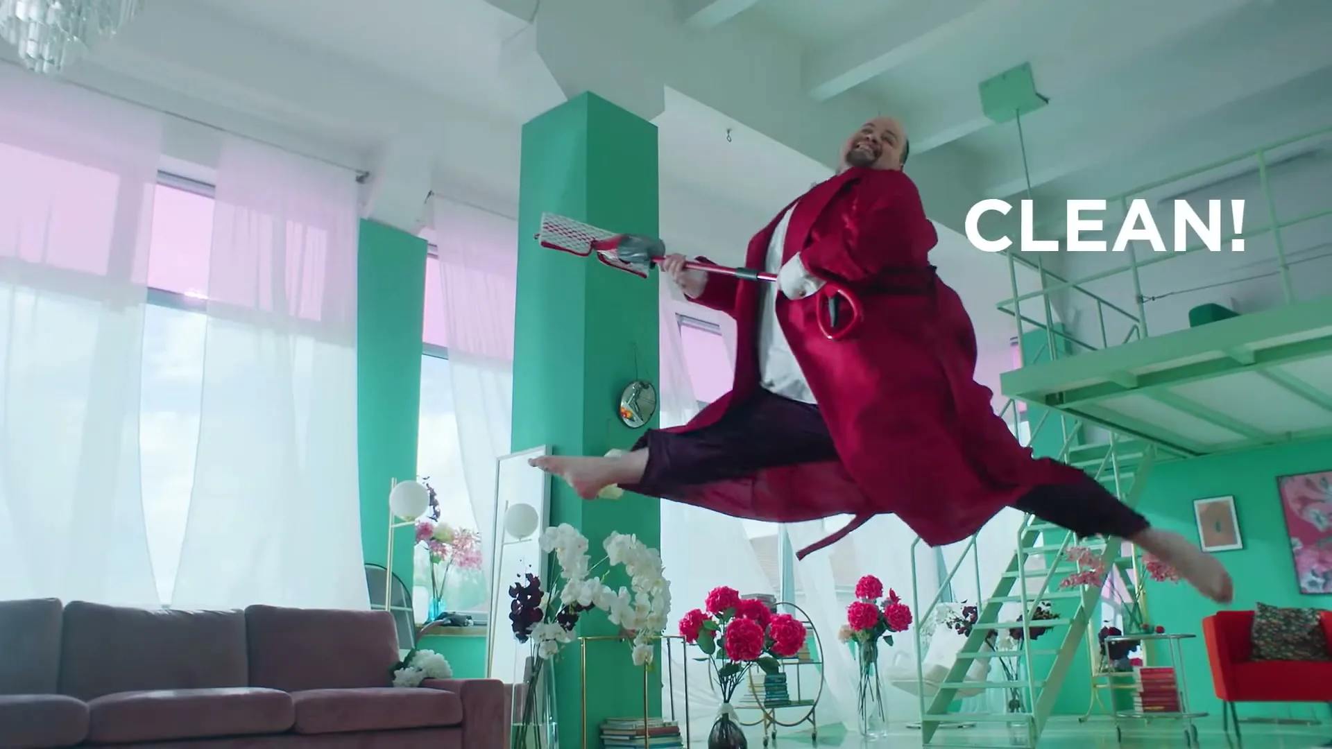 A man in a red robe and purple pants leaps in the air, holding a mop like a guitar, in a brightly lit, colorful living room.