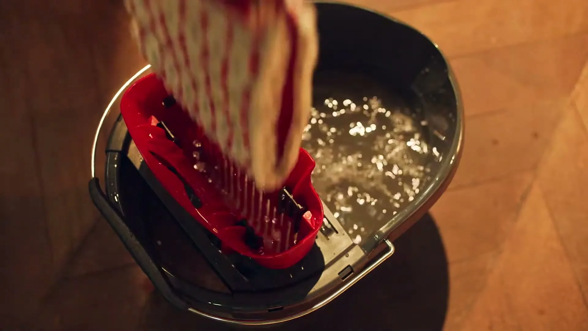 A red mop head is being wrung out in a black bucket with dirty water, seen from above on a wooden floor.