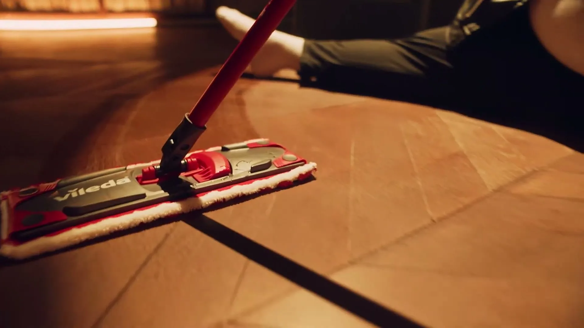 A Vileda mop is seen on a wooden floor, with a person's leg and foot visible in the background, in a dimly lit room.