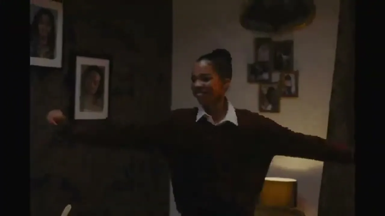 A young Black woman in a school uniform dances playfully, holding a frying pan like a guitar in a dimly lit living room.