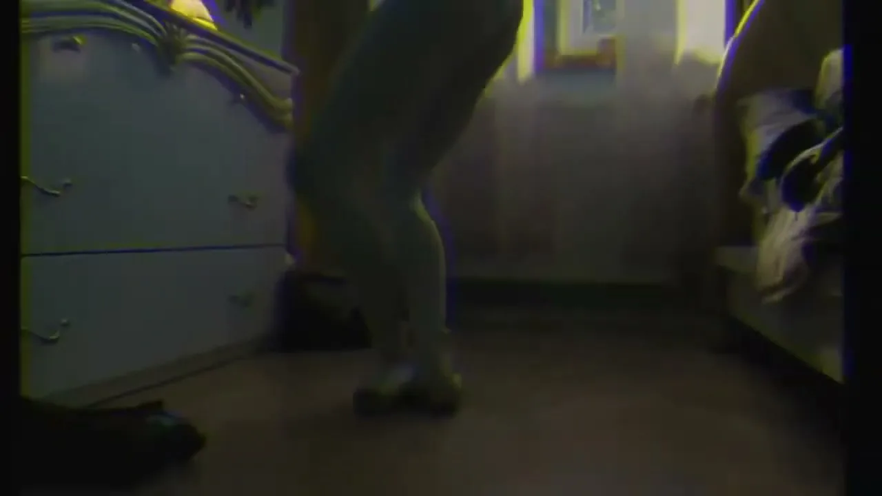 A young person in a light blue leotard and ballet shoes practices ballet in a dimly lit bedroom.