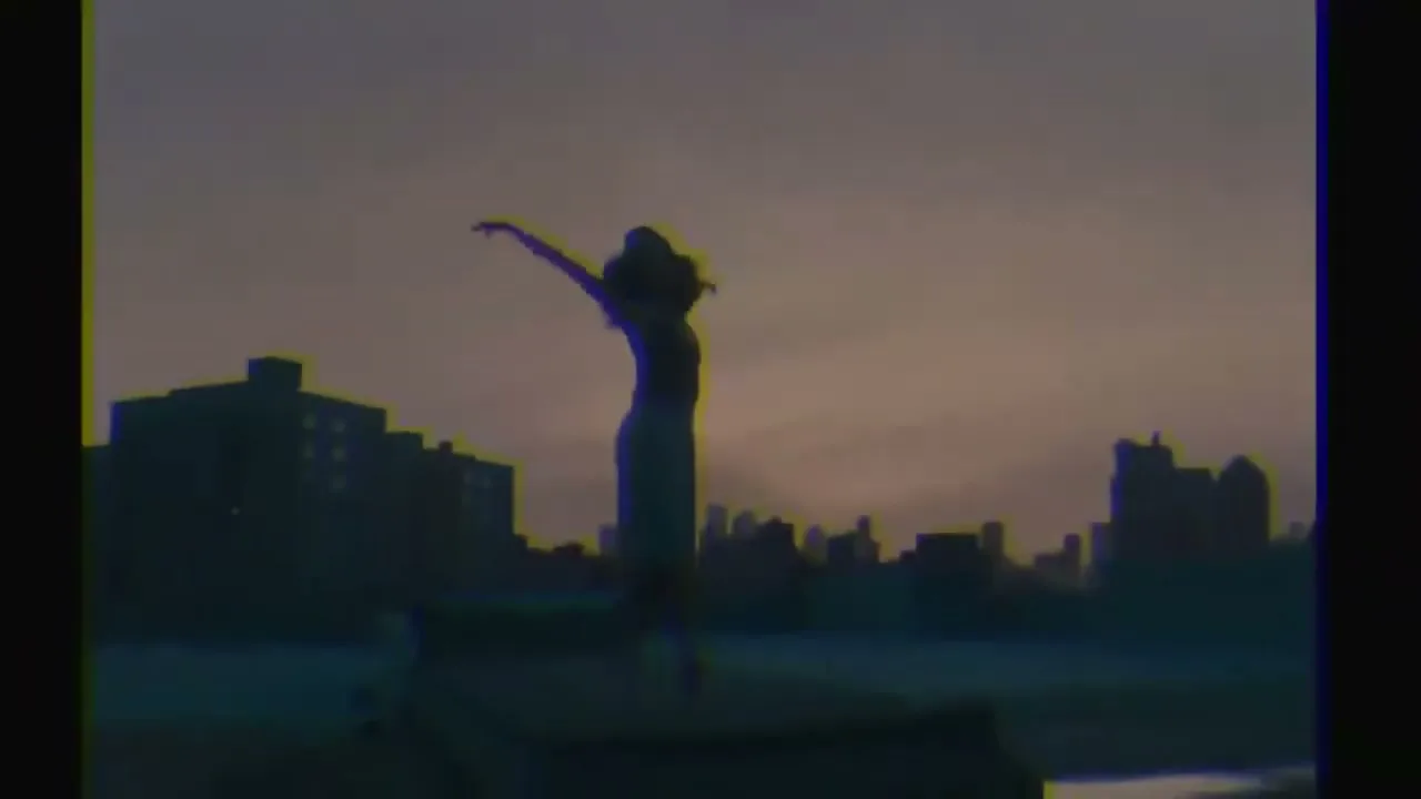 A young woman in silhouette dances with arms outstretched on a rooftop at dusk, with city buildings in the background.