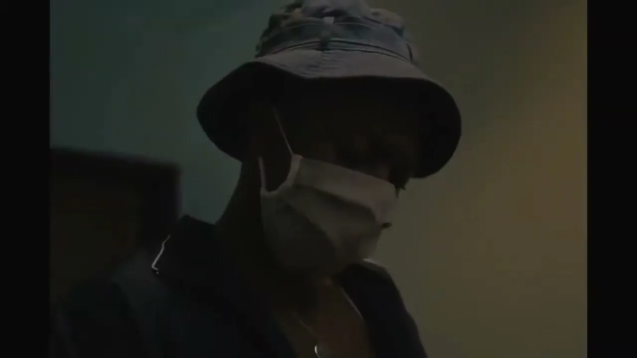 A young man in a bucket hat and face mask looks down, standing in a dimly lit room.