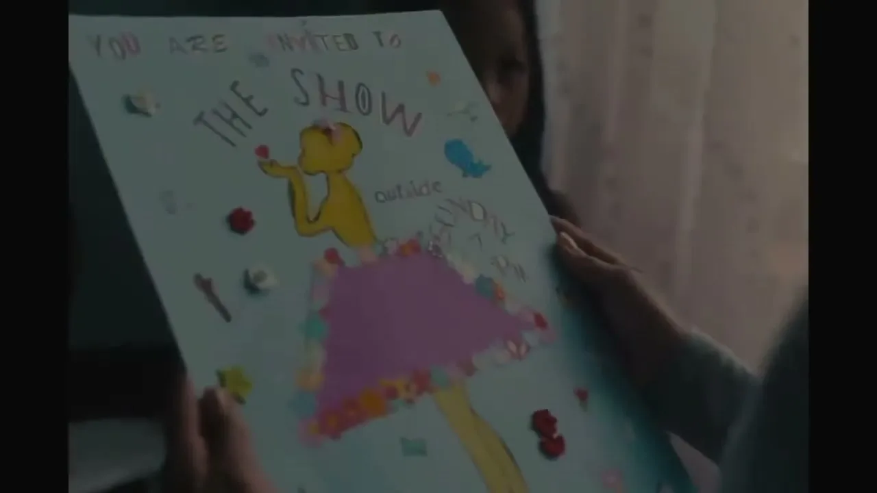 A child's hands hold up a handmade invitation for "The Show" with a drawing of a dancer, in a dimly lit room.
