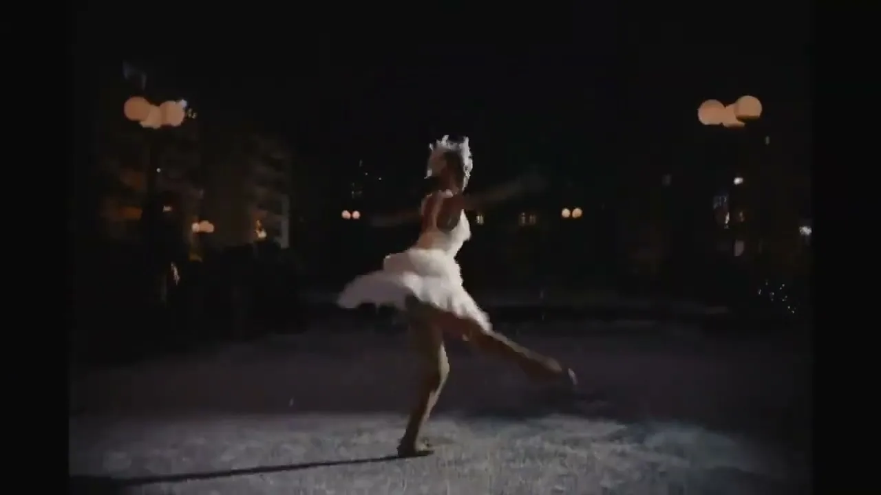 A young female ballerina in a white tutu dances gracefully on a dark street at night, with streetlights visible in the background.