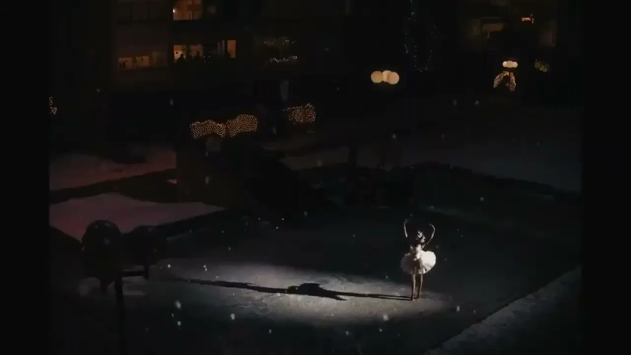 A young girl in a white tutu performs ballet in a spotlight on a snowy street at night, with festive lights in the background.