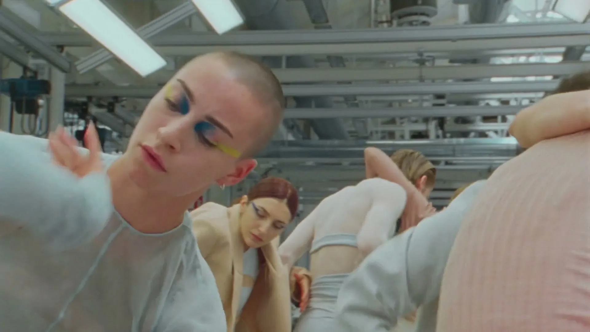 A group of models with avant-garde makeup and neutral clothing pose and move in a factory setting.