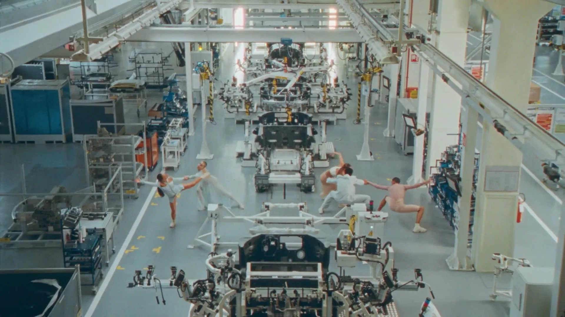 A group of dancers in leotards perform ballet moves in a car manufacturing plant, surrounded by car frames and machinery.