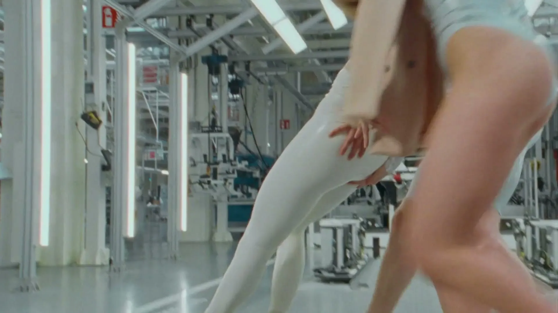 Two women in futuristic bodysuits and a beige coat dance in a factory with a car chassis in the background.