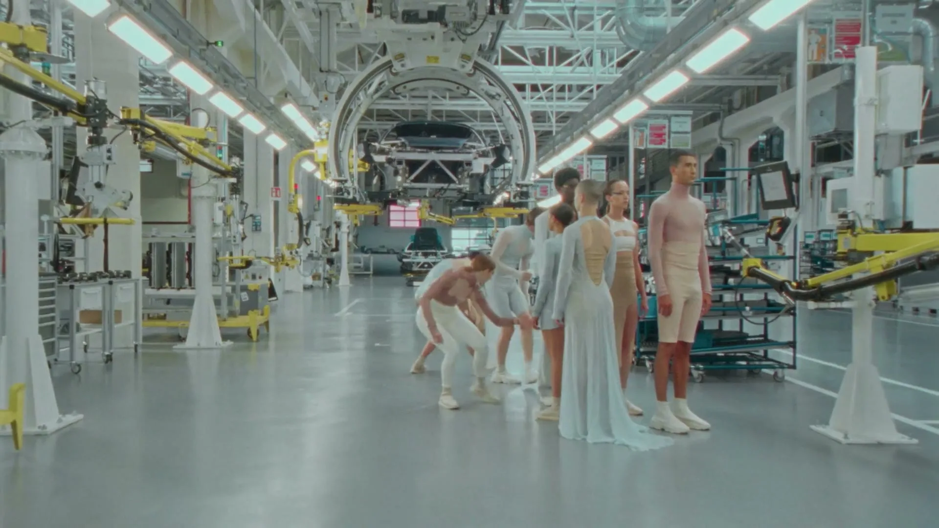 A group of models in various light-colored outfits pose and move in a futuristic car manufacturing plant.