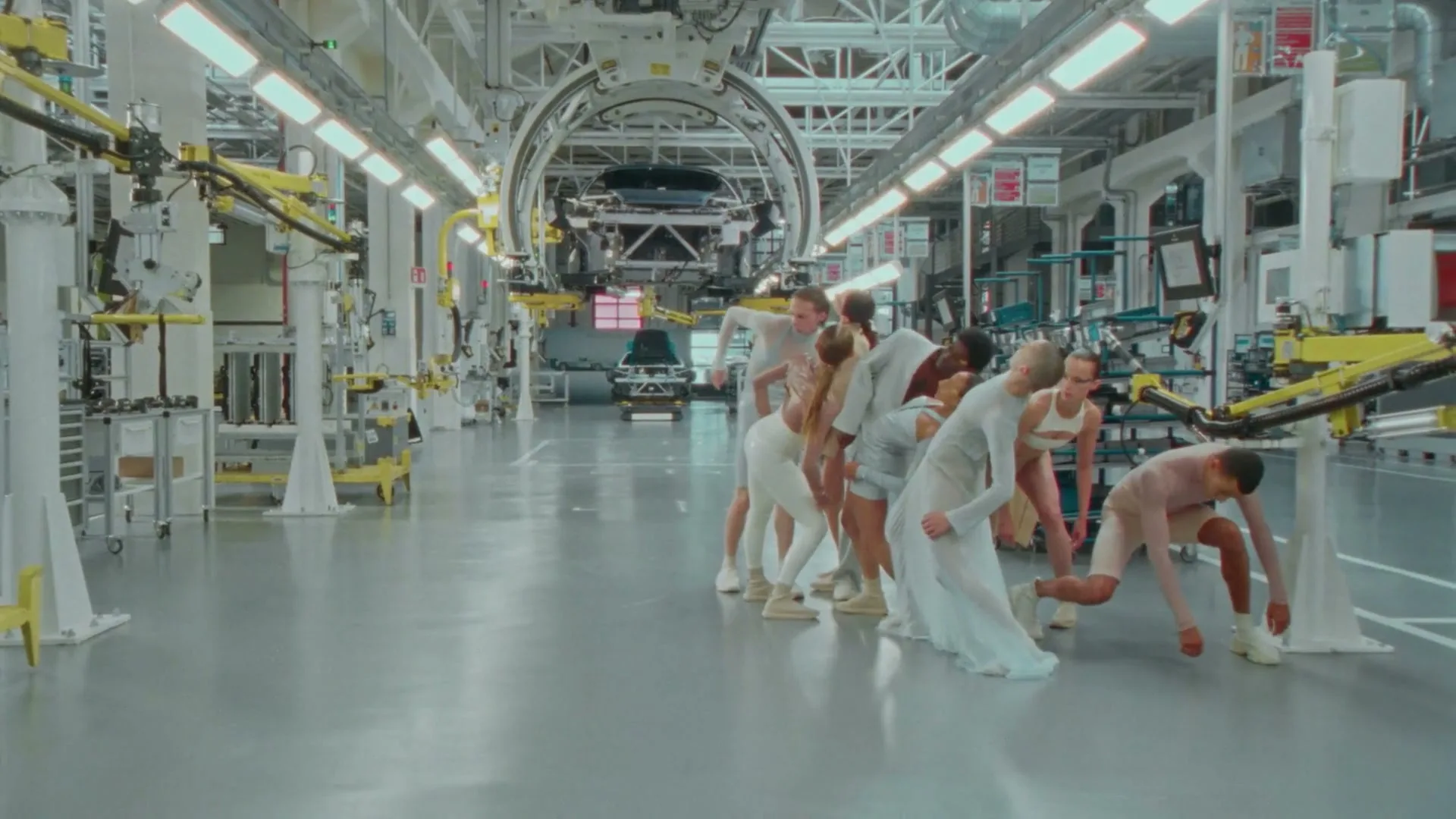A group of dancers in light-colored, flowing costumes perform a synchronized, fluid movement in a brightly lit, modern car factory.