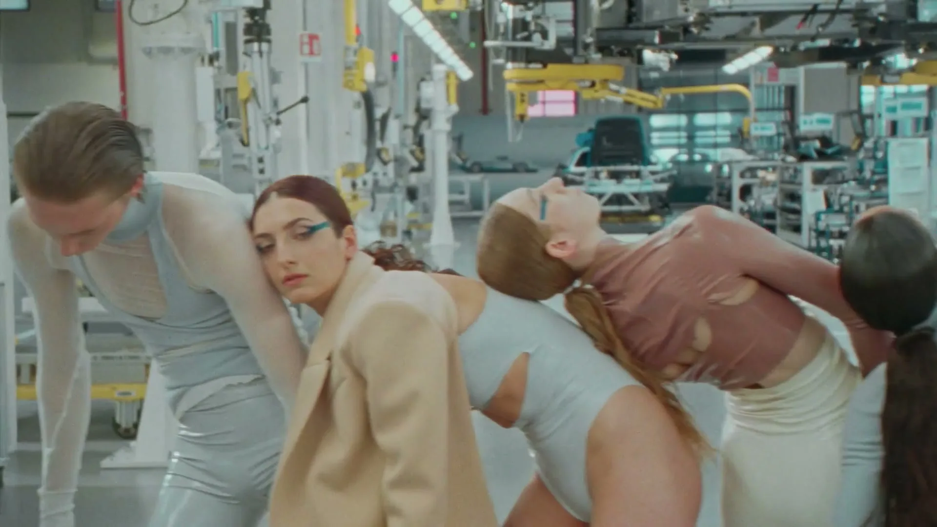 A group of models in futuristic outfits pose in a modern factory setting, leaning on each other in a line.