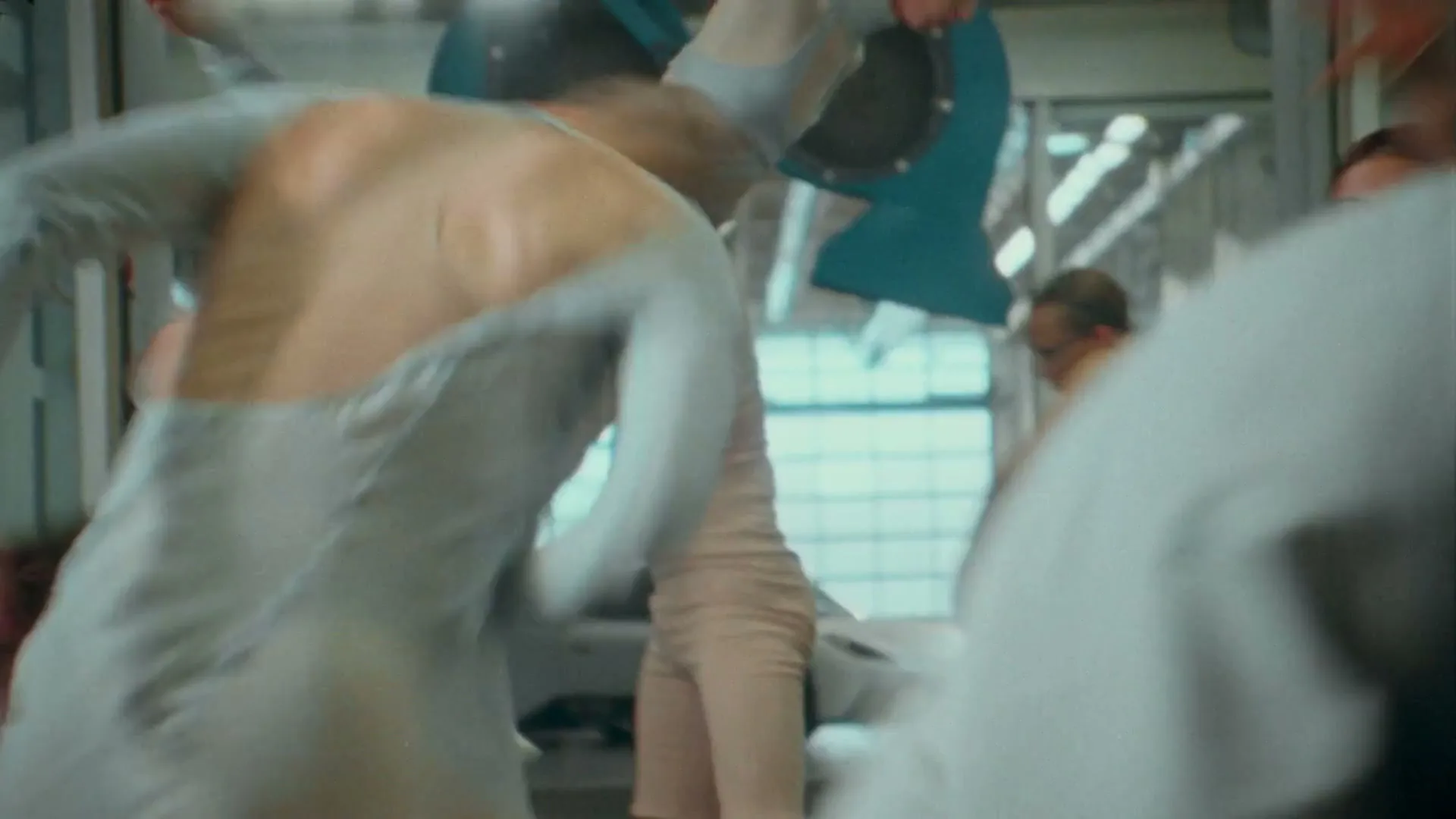 A group of dancers in skin-tight, light-colored outfits move chaotically in a brightly lit, industrial-looking space, with one dancer carrying another over their shoulder.