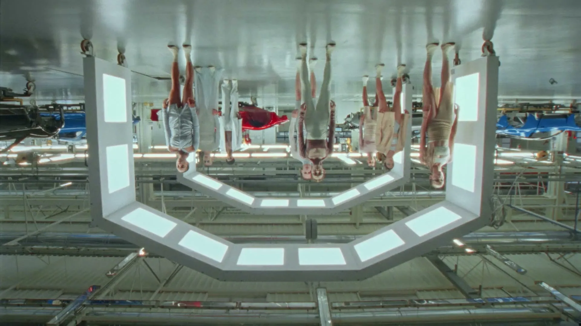 Several people are seen upside down, suspended from the ceiling of a factory, wearing various outfits, as if in a fashion show.