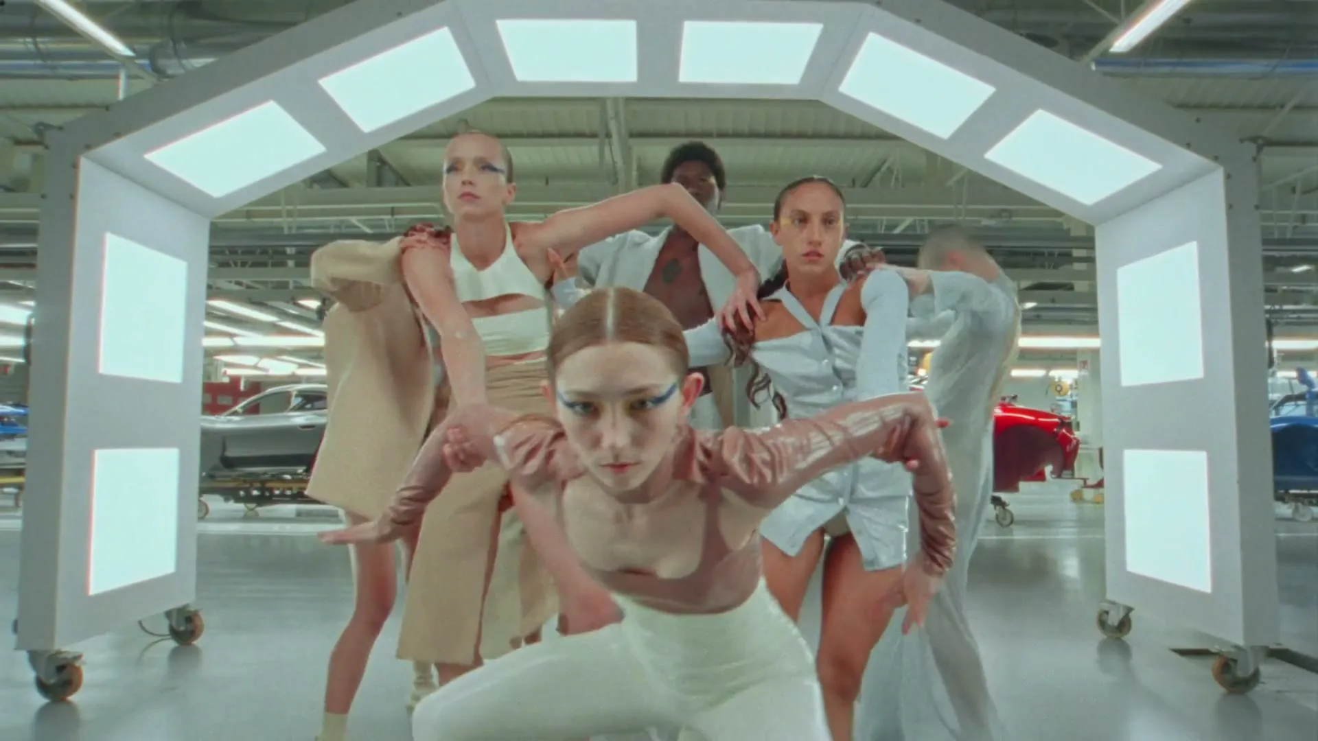 A group of six dancers in futuristic, light-colored outfits perform synchronized movements under a lit archway in a car manufacturing plant.