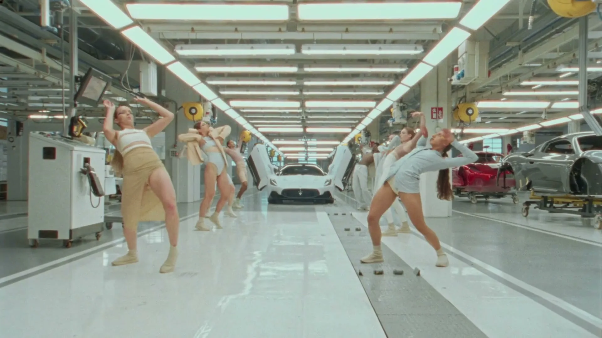A group of dancers in neutral-toned outfits perform synchronized movements in a brightly lit, futuristic car manufacturing plant, with a white Maserati MC20 visible in the background.