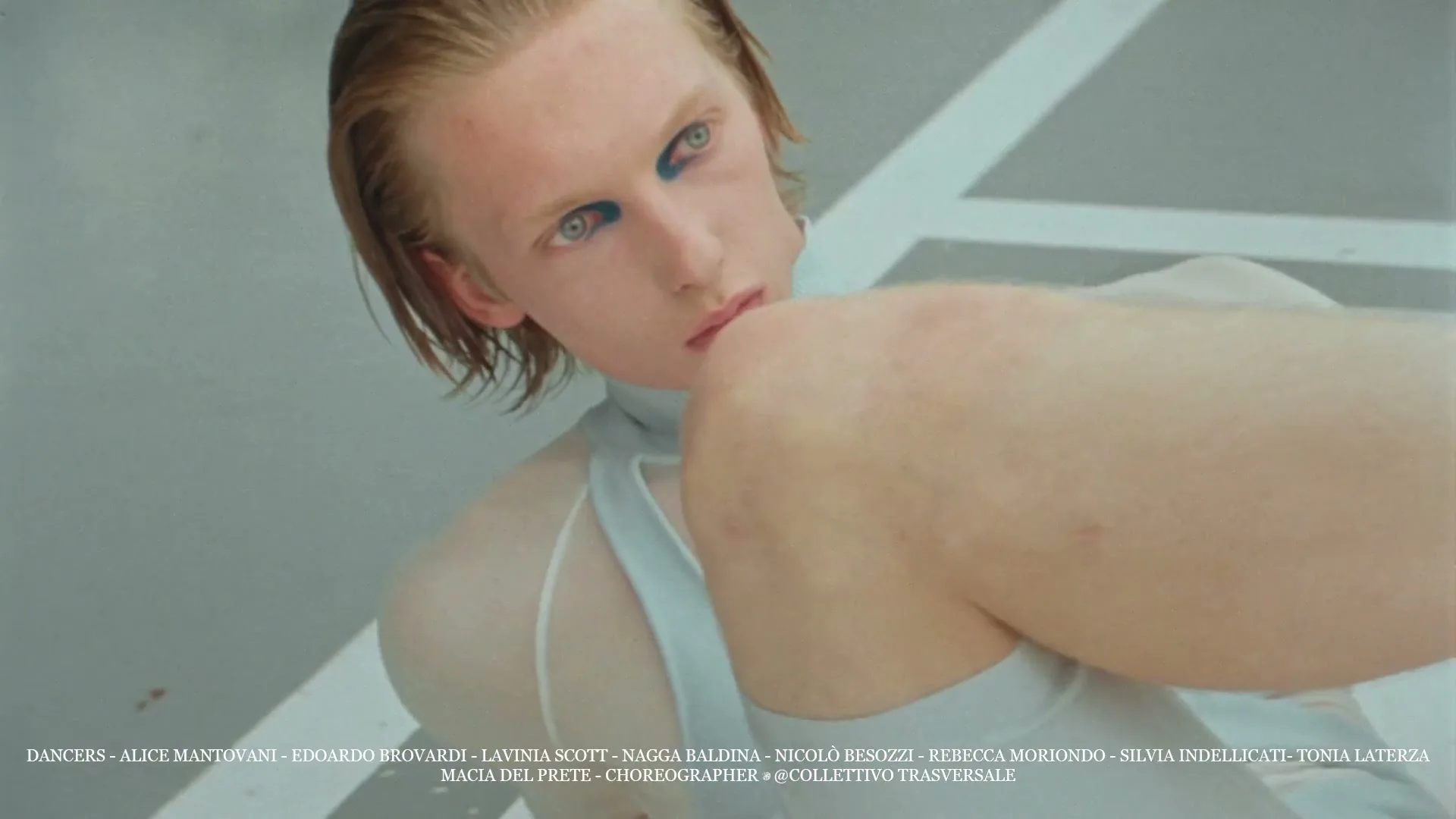 A young person with blue eyeliner and wet hair looks intensely at the camera while moving their arm in a white, minimalist setting.