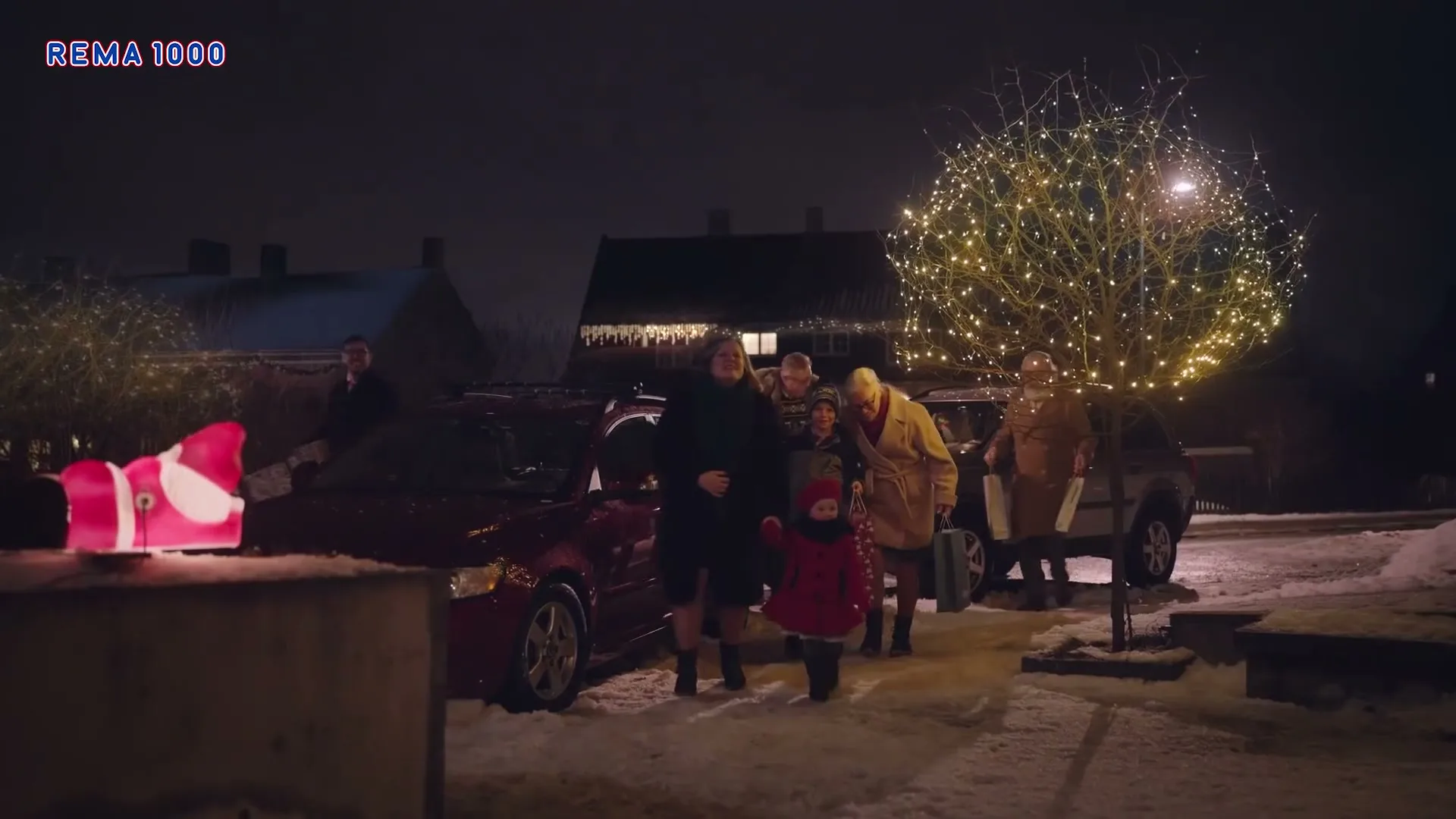 A family, including two adults and two children, arrives home at night, carrying groceries and gifts, with a glowing Santa decoration in the foreground.