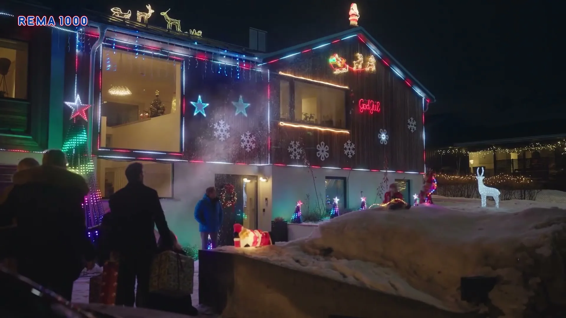 A wide shot of a house covered in colorful Christmas lights and decorations, with several people standing outside in the snow at night.