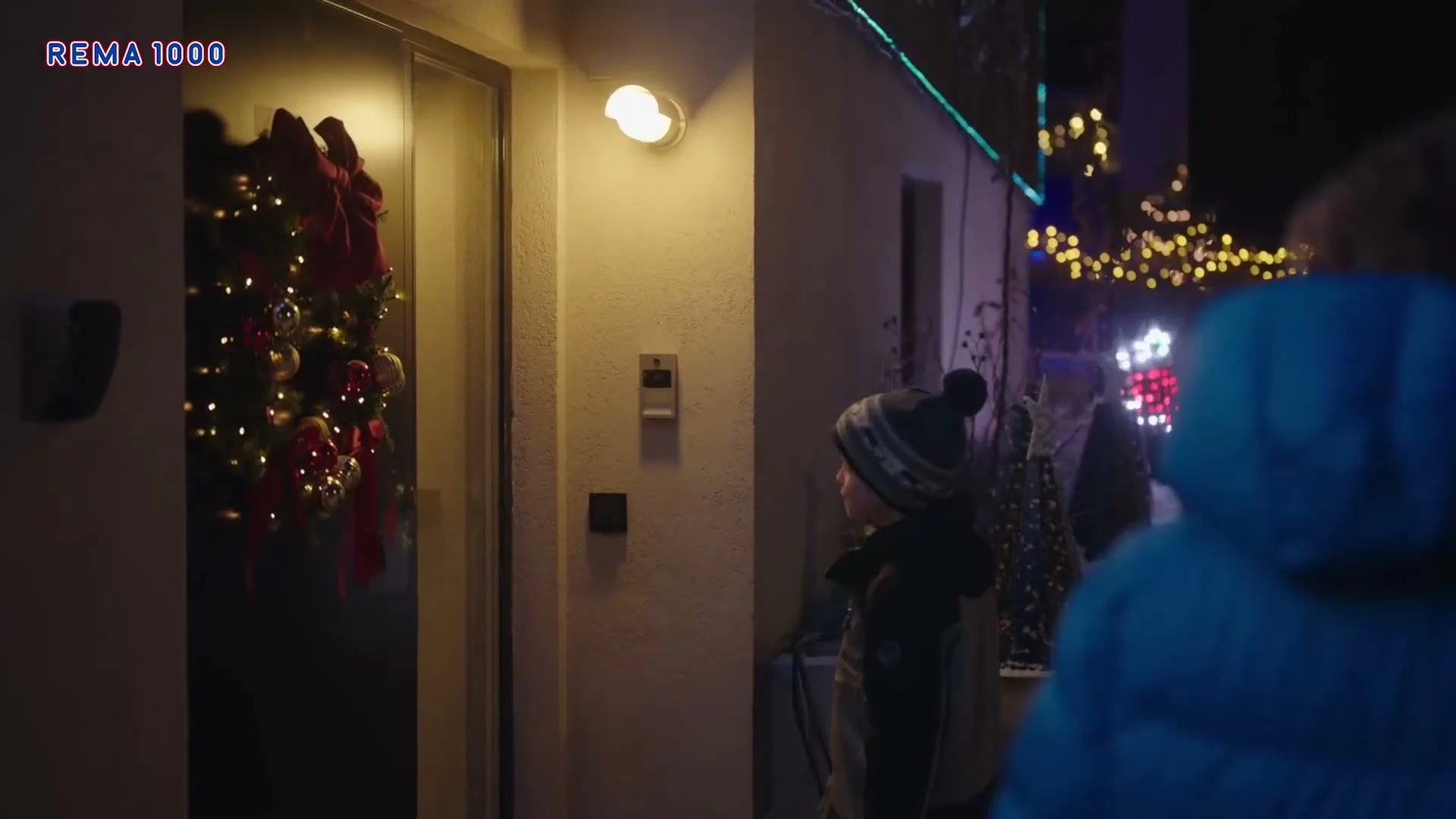 A young boy in winter clothes stands by a door decorated with a Christmas wreath, looking back at someone off-screen at night.