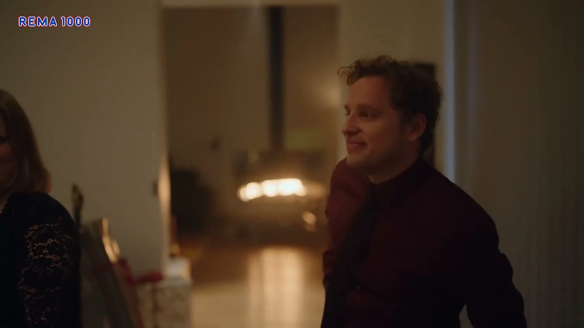 A man in his 30s, wearing a maroon shirt and black tie, walks past a woman in a dark room, smiling and looking to his right.