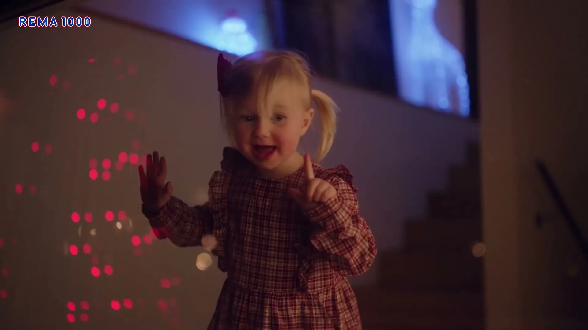 A cute blonde toddler girl in a plaid dress with pigtails and a red bow smiles and points, standing by a glass wall with red lights.