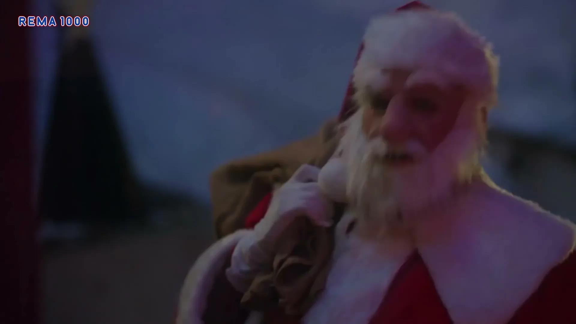 Santa Claus, dressed in a red suit with a white beard, carries a sack over his shoulder, looking around in a festive outdoor setting at night.