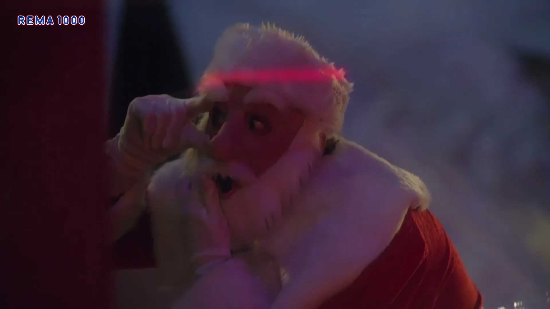 Santa Claus, dressed in a red suit with white trim, peeks around a corner with a surprised expression, illuminated by green and red lights.