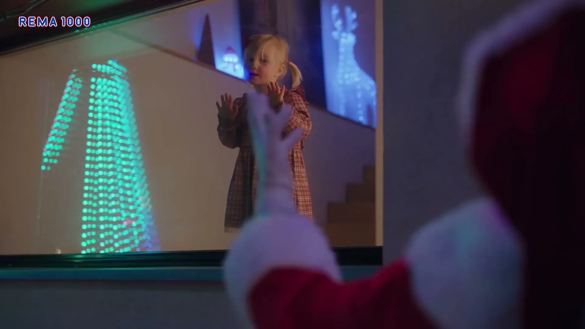 A young blonde girl in a plaid dress looks out a window at Santa Claus, who is partially visible in the foreground, at night.