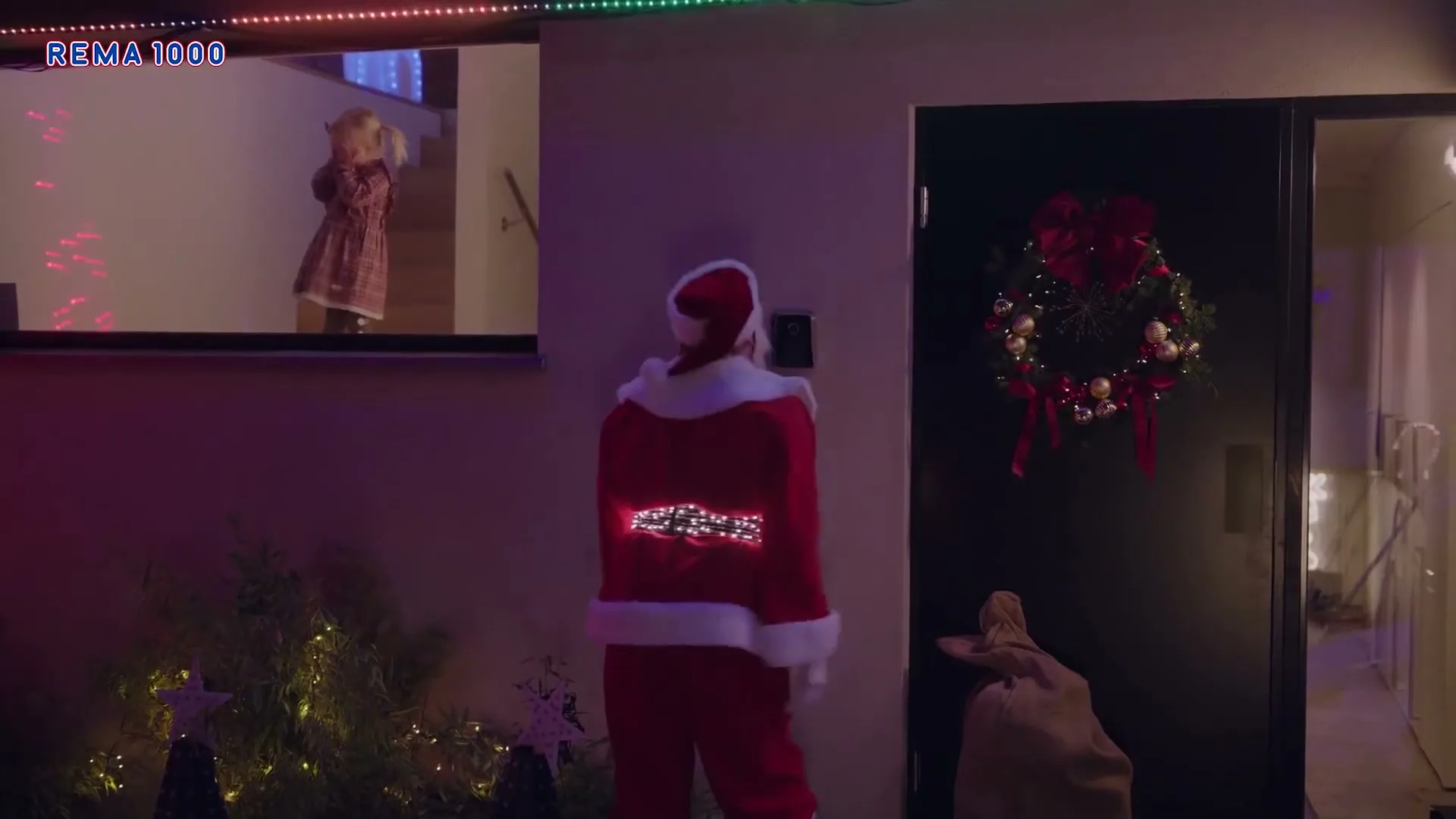 A person dressed as Santa Claus rings a doorbell while a young girl in a dress watches from an upstairs window, at night.