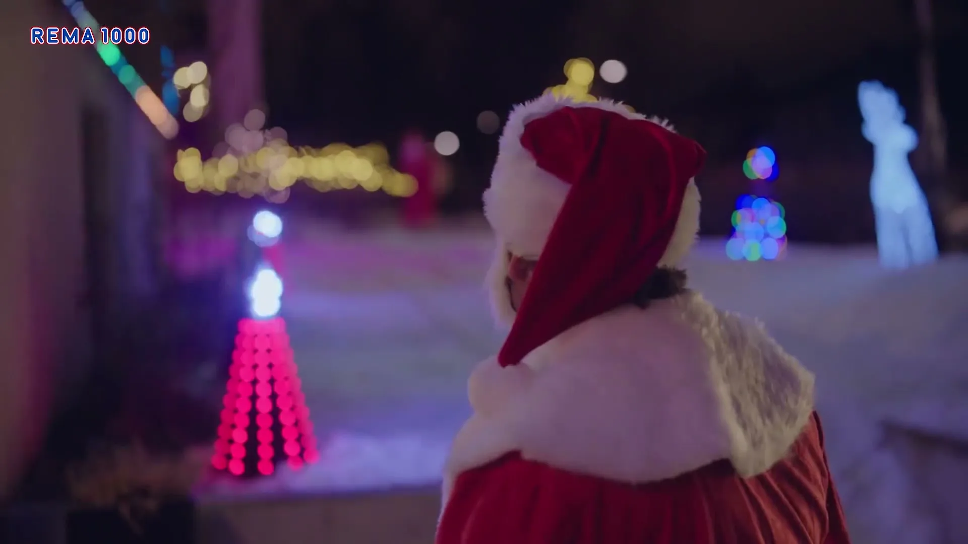 A man dressed as Santa Claus, wearing a red suit and white beard, looks over his shoulder at another Santa in a snowy, decorated yard at night.