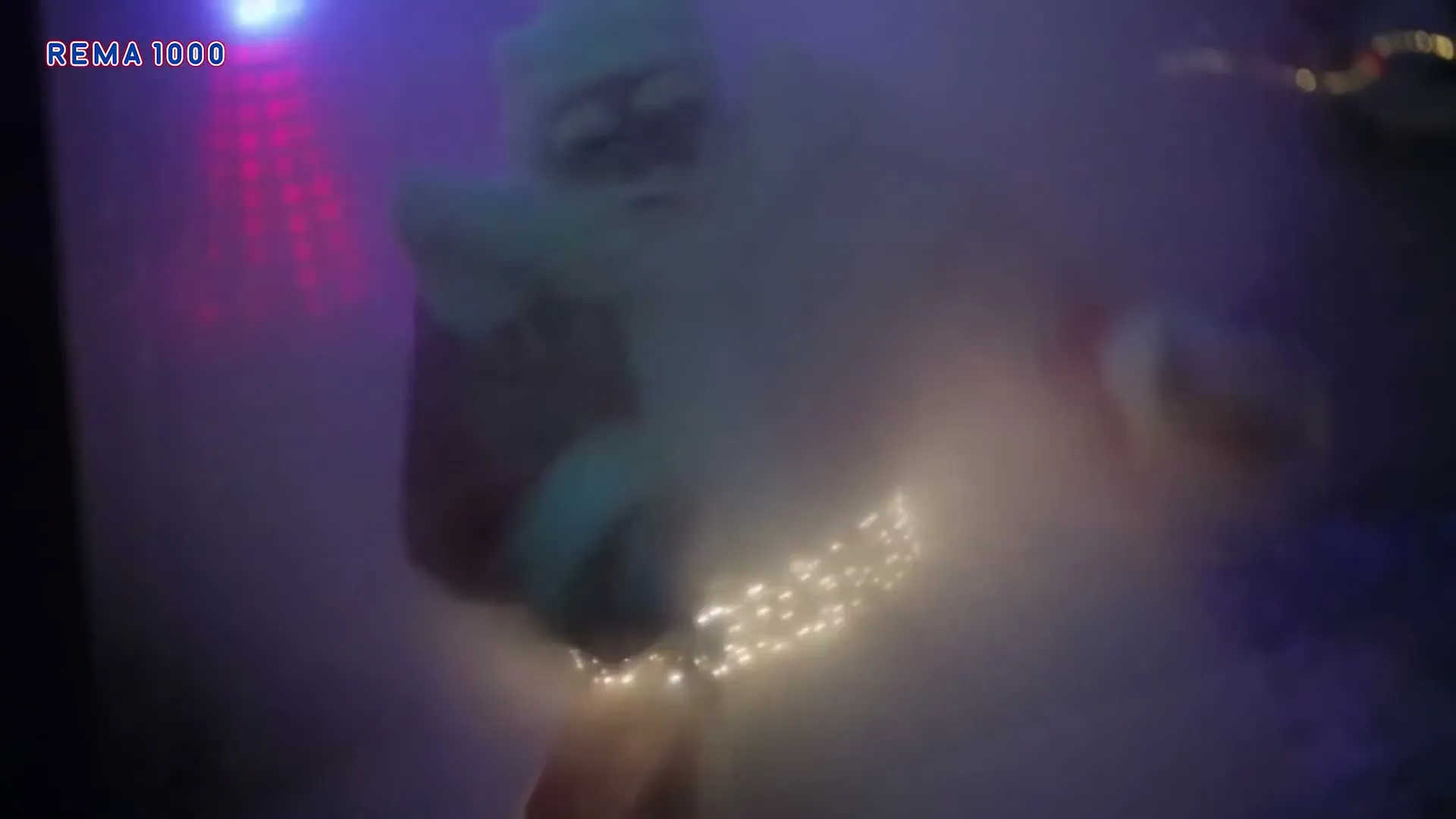 A blurry Santa Claus, wearing a red suit with glowing lights around his waist, dances in a smoky, purple-lit room.