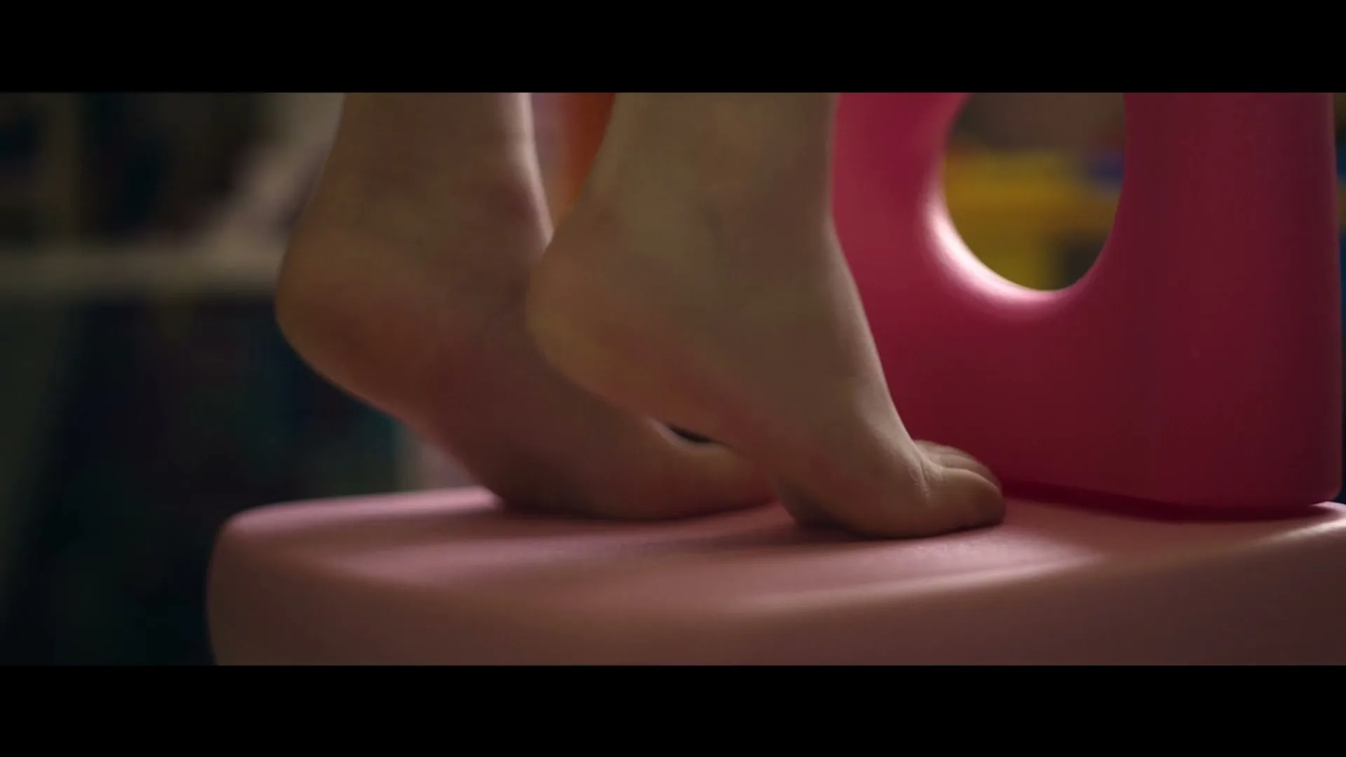 A child's bare feet stand on a pink plastic stool, stretching upwards, in a dimly lit room.