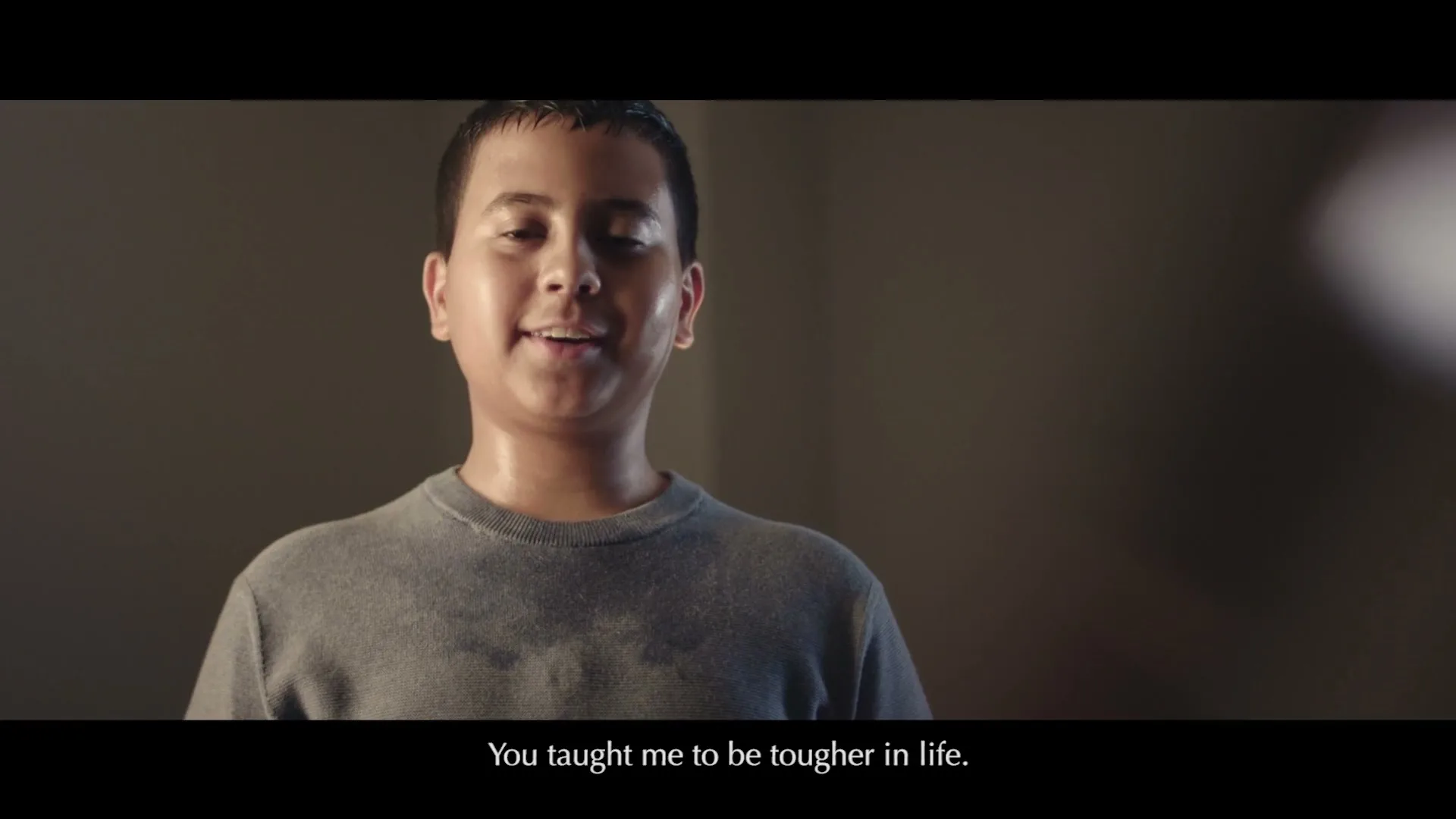 A young boy, sweaty and wearing a grey t-shirt, smiles at the camera in a dimly lit room.