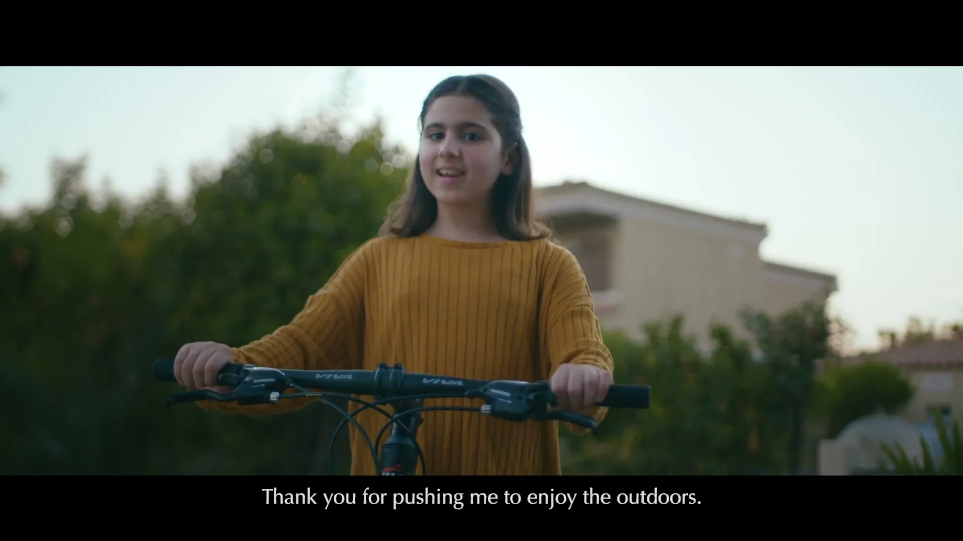 A young girl with long dark hair, wearing a mustard yellow sweater, holds the handlebars of a bicycle and speaks to the camera in a suburban neighborhood.