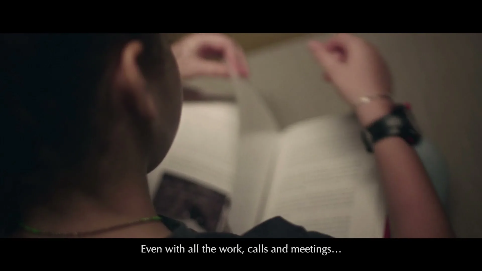 A person with dark hair and a black t-shirt reads a book, turning pages, in a dimly lit room.