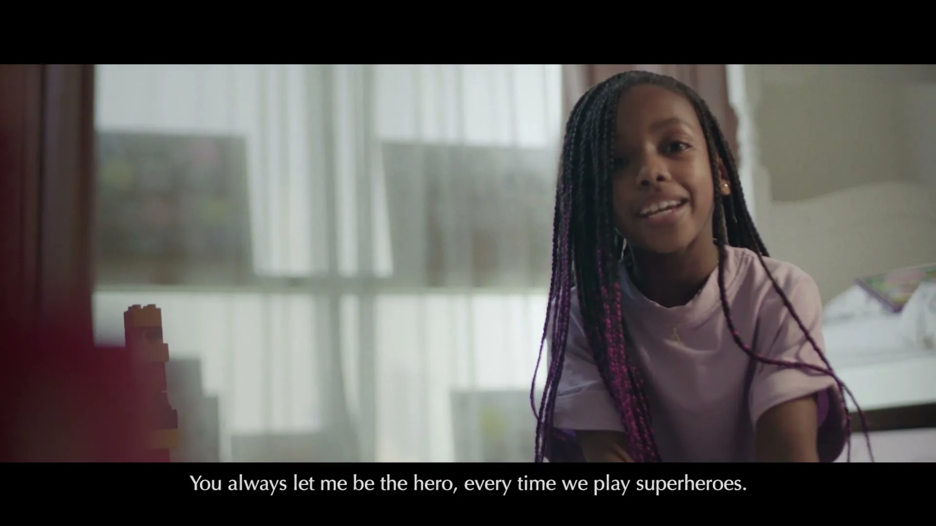 A young girl with long braids and a light purple t-shirt looks directly at the camera, smiling, in a softly lit room.