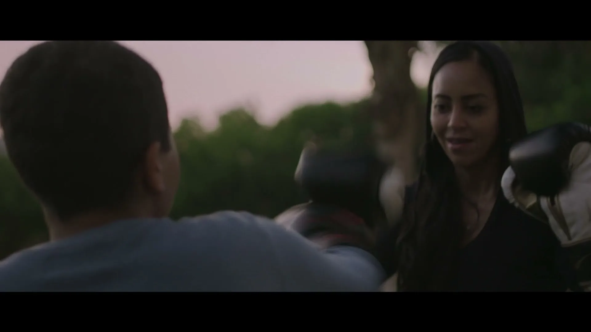 A young boy in a blue t-shirt practices boxing with a woman in a black hoodie and boxing gloves, outdoors at dusk.