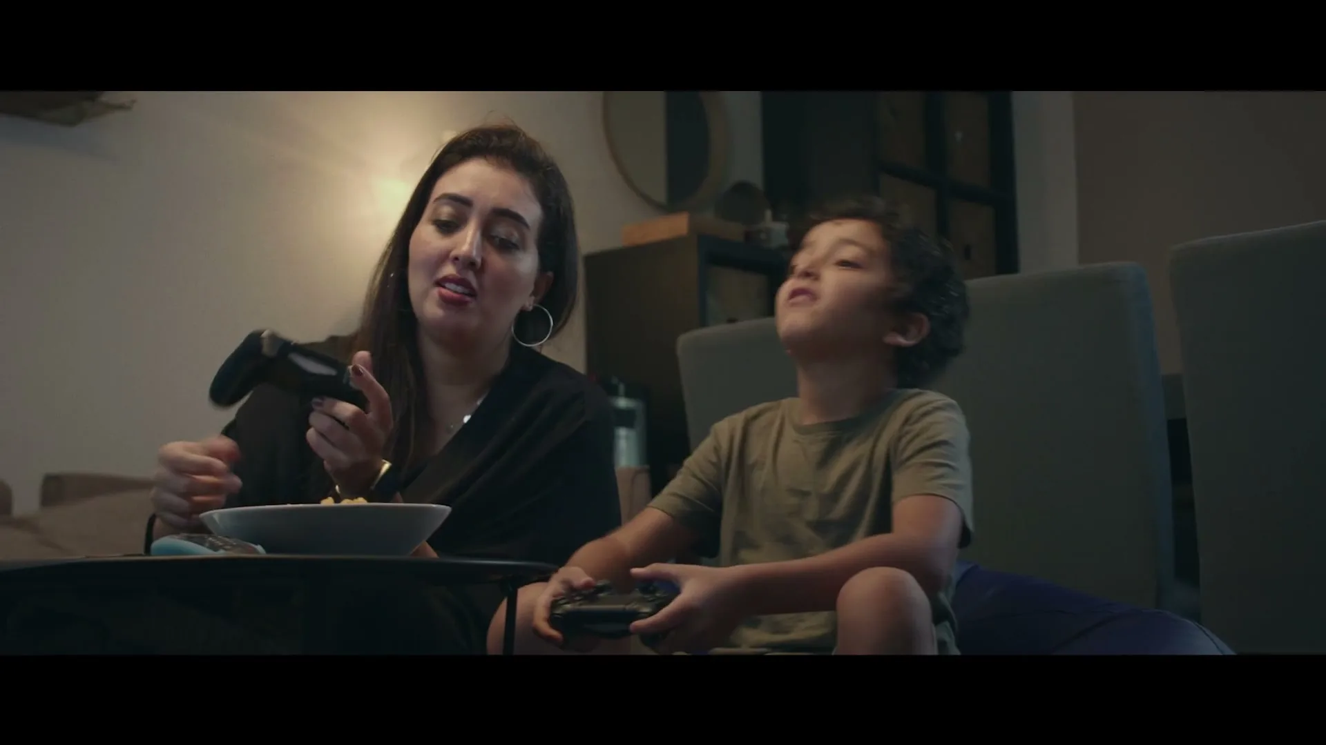 A woman and a young boy sit on a couch, playing video games and eating snacks in a dimly lit living room.