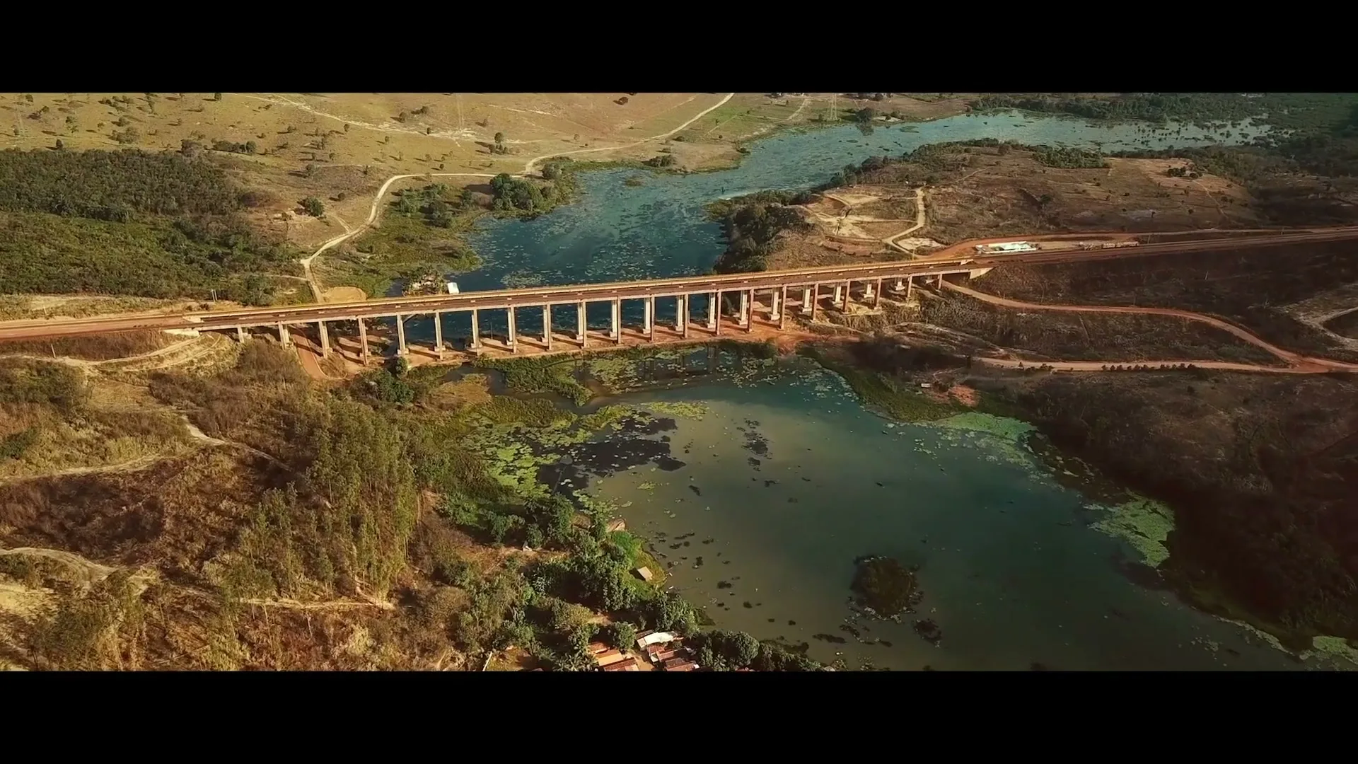 An aerial view shows a long bridge over a river, surrounded by dry, hilly terrain and a small cluster of houses, under a bright sky.