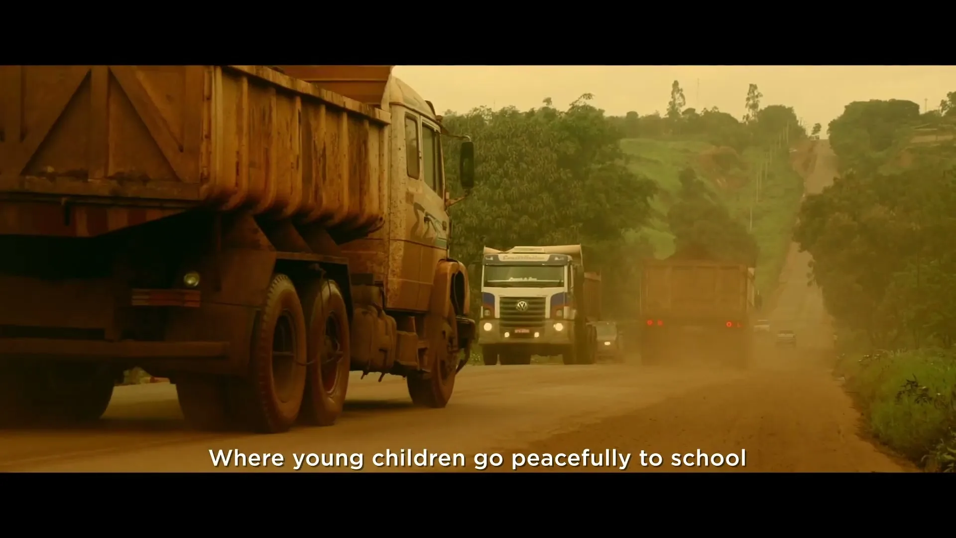 A line of large, dirty dump trucks drives down a dusty, unpaved road, kicking up clouds of dust in a rural, hilly landscape.